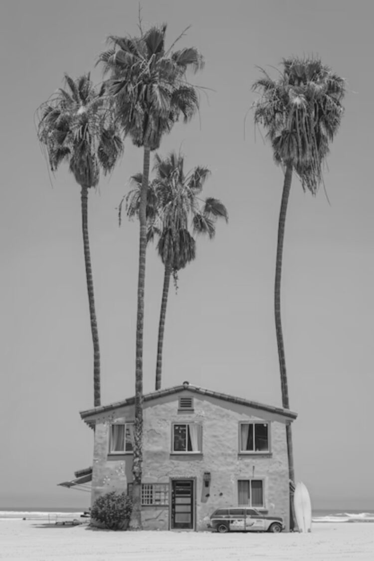A small beach house with palm trees towering above it, a surfboard leaning against the wall, and an old car parked outside.